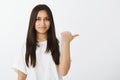 Waist-up shot of timid unconfident good-looking girl with dark hair, pointing right with thumb and smiling casually