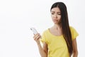Waist-up shot of unhappy upset beautiful young woman without makeup in yellow t-shirt holding smartphone looking at Royalty Free Stock Photo