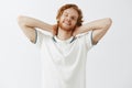 Waist-up shto of lazy carefree happy redhead male model with beard in white polo shirt holding hands behind head and