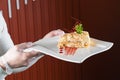 A waiter in gloves holds a white plate of napoleon cake decorated with a spiral of red chocolate, mint and berry jam on Royalty Free Stock Photo