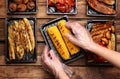 Waiter in gloves putting grilled corn cobs into containers at wooden table, top view. Food delivery service