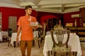 Waiter in the uniform walking with glasses on the tray in the restaurant at the tropical island