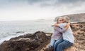 Waiting for the sunrise. Senior couple with grey hair sitting on the rocks. Clear sky and ocean. Embraced with love