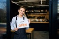 Waitress wearing apron standing at restaurant entrance. Woman attend new customers in her coffee shop. Happy woman showing open