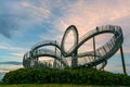 Walkable Tiger & Turtle roller coaster sculpture on Magic Mountain with some people in motion blur against a cloudy sunset sky,