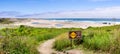 Walking path going through green shrubs towards a sandy beach; sign warning of Recurring Rip Currents posted on the trail; Gazos