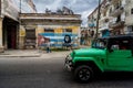 A wall with the Cuban flag a man walking next and a green jeep crossing in front