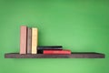 On the wall of green color there is a dark wooden shelf with a texture on which several books stand vertically from the left and