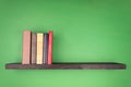 On the wall of green color there is a dark wooden shelf where a group of books stand vertically from the left