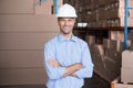 Man standing with arms crossed in warehouse among stacked boxes, wearing white hard hat Royalty Free Stock Photo