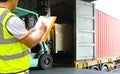 Warehouse worker holding clipboards control forklift loading shipment goods into container truck.