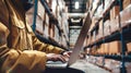 A warehouse worker sits in the aisle of a large warehouse, using a laptop to manage inventory, with rows of stacked boxes behind