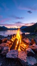 Warm campfire burns beside a serene lake at twilight with colorful sky and mountains Royalty Free Stock Photo