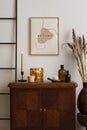 Warm and cosy living room interior with mock up poster frame, wooden sideboard, gold candle, orange vase with dried flowers, glass