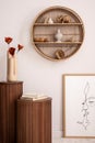 Warm and cozy composition of living room interior with round shelf , beige pouf, mock up poster frame, vase with dried flowers and
