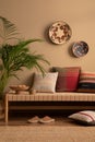 Warm and cozy ethno living room interior with couch, patterned pillows, plants, flowerpots, basket on wall, slippers, wooden bowl