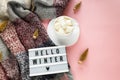 Warm, cozy winter scarf and white cup of coffee with white marshmallow as frame on pastel pink background.