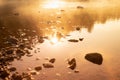 Warmy golden sunlight on quiet water surface of lake, orange haze, ripple sun glares, blinks, reflection, stones on shore in early
