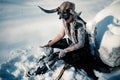 Warrior woman in image of viking with horned helmet and ax sits on snow next to big stone