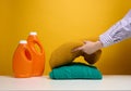 Washed folded clothes and plastic orange large bottles with liquid detergent stand on a white table, yellow background. Routine Royalty Free Stock Photo