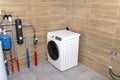 A washing machine standing in the corner of a modern gas boiler room, lined with ceramic tiles imitating wood, visible water pipes Royalty Free Stock Photo