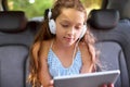Watching her movies in the backseat. a young girl sitting in a car backseat wearing headphones and using a digital