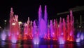 A water feature in the city is illuminated with purple and red lights at night