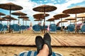 Water shoes feet on a sunbed facing beach, rush umbrellas, wood pontoon and white cloud sky Royalty Free Stock Photo