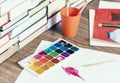 Watercolor paint, brushes well used and sheet of paper on wooden table with stack of books background Royalty Free Stock Photo