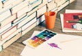 Watercolor paint, brushes well used and sheet of paper on wooden table with stack of books background Royalty Free Stock Photo