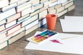 Watercolor paint, brushes well used and sheet of paper on wooden table with stack of books background Royalty Free Stock Photo