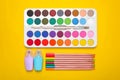 Watercolor palette, colorful pencils and markers on yellow background, flat lay