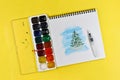 Watercolors paints and drawing young green Christmas tree covered with fluffy snow, album on yellow paper background Royalty Free Stock Photo