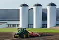 John Deere 8R 310 tractor pulling a Poettinger cultivator with a barn and silos in the background