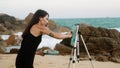 Artist paints turbulent sea on canvas at rocky shore. Waves crash behind as woman with palette creates art. Hobbyist in