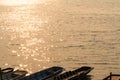 Wavy water surface with glittering orange sun light and row of long-tailed boats parking on the shore.