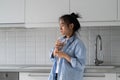 Weakened lonely Asian woman standing in kitchen of own home with glass in hands with eyes closed