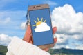 Weather forecast concept with hand holding mobile phone with icon of cloud covering sun in front of blue sky