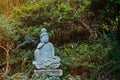 A weathered stone statue of a Buddha or Buddhist monk sitting and observing world, meditates under green trees in japanese garden