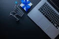 Web shopping. Laptop computer, shopping trolley and white gift with blue ribbon on dark background. Ecommerce and Royalty Free Stock Photo