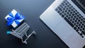 Web shopping. Laptop computer, shopping trolley and white gift with blue ribbon on dark background. Ecommerce and Royalty Free Stock Photo