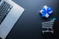 Web shopping. Laptop computer, shopping trolley and white gift with blue ribbon on dark background. Ecommerce and Royalty Free Stock Photo