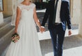 Wedding blonde couple is walking in old city. Bride in lace satin dress and groom are holding hands together in the street with