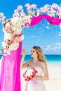 Wedding ceremony on a tropical beach in purple. Happy blond bride with wedding bouquet under the arch decorated with flowers on t Royalty Free Stock Photo