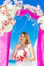 Wedding ceremony on a tropical beach in purple. Happy blond bride with wedding bouquet under the arch decorated with flowers on t Royalty Free Stock Photo