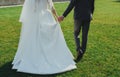 Wedding couple is holding hands and walking outside in the bright green lawn. Bride in white elegant wedding dress with train is Royalty Free Stock Photo