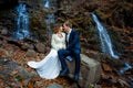 Wedding couple sitting on stone and lovingly look at each other. Waterfall background. Royalty Free Stock Photo