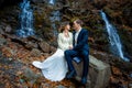 Wedding couple sitting on stone and lovingly look at each other. Waterfall background Royalty Free Stock Photo
