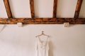 Wedding dress hangs on a hanger on a white wall under wooden ceiling beams