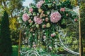 Wedding glass arch with white patterns decorations is standing outdoors in the green summer park. Pink hortensia and roses flowers